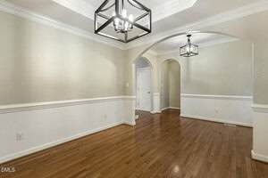 A dining room with hardwood floors, neutral beige walls, white trim, and wainscoting. Two black geometric light fixtures hang from the ceiling, and arched openings lead to an adjacent hallway and another room.