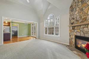A spacious living room with light-colored carpet, a stone fireplace, and large windows. The room opens to a kitchen with wood cabinets and a green accent wall, with hardwood flooring in the adjoining area.