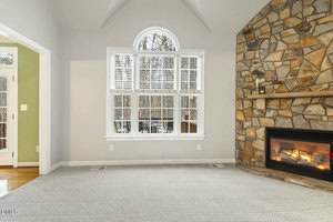 A living room with light gray carpet, a large window with white blinds, and a stone fireplace with a lit fire. A small wooden shelf above the fireplace holds a candle and small decor items. Pale walls and a high ceiling.