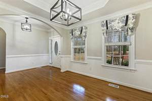A bright entryway or dining area with hardwood floors, white walls, crown molding, modern light fixtures, and two windows with patterned valances looking out onto a yard with trees and a wooden bench.