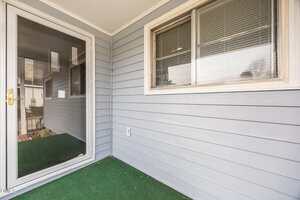 A small covered porch area with light gray siding, a large window with blinds, a glass storm door with a brass handle, and green indoor-outdoor carpet on the floor.