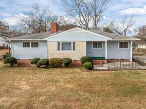 A single-story house with light blue siding, a brick chimney, and stone accents around the front window. Neatly trimmed bushes line the front, and the yard has brownish grass. Leafless trees stand in the background.