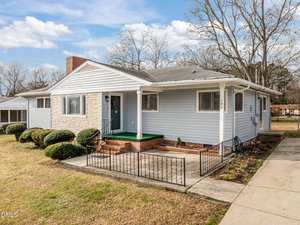 Charming single-story home at 109 Warren Avenue, Oxford, with light blue siding, a brick chimney, and a covered front porch featuring green flooring. Enjoy trimmed bushes, a small metal railing, concrete driveway, and scenic trees under a clear sky.