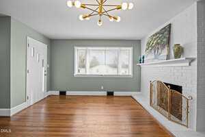 A bright living room with hardwood floors, a modern chandelier, a large window, a white painted brick fireplace with a decorative screen, and a landscape painting on the mantel.
