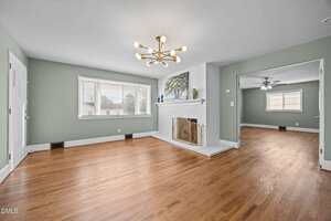 A spacious living room with light green walls, large window, hardwood floors, a white brick fireplace with a gold screen, and a modern chandelier. An adjacent room with a ceiling fan is visible through a wide doorway.