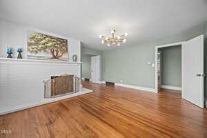 A spacious living room with hardwood floors, a white brick fireplace, green walls, a modern gold chandelier, and a painting and candles on the mantel. The room has an open doorway and a partially open white door.