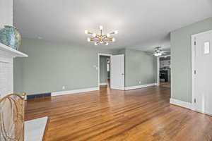 A spacious, empty living room with light green walls, hardwood floors, a modern chandelier, and a white brick fireplace. An open doorway leads to a kitchen with dark cabinets and a ceiling fan.