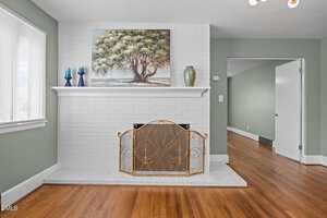 A white brick fireplace with a decorative gold screen, a mantel holding three vases and a painting of a tree. The room has light green walls and hardwood floors. An open doorway leads to an adjacent room.