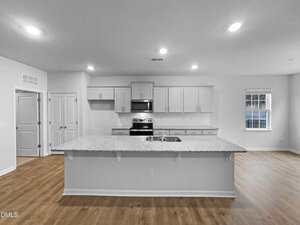 A modern kitchen with white cabinets, stainless steel appliances, and a large granite island with a sink. Hardwood floors and recessed lighting are visible, with a window and doors in the background.