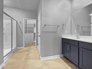 A modern bathroom with light-colored walls and flooring, featuring a glass shower, a white sink with dark cabinetry, two towel racks, and a walk-in closet with built-in shelves and drawers in the background.
