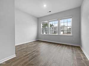 Empty room with light gray walls, wood-style flooring, and three large windows letting in natural light. The windows overlook a residential area with neighboring houses visible outside.