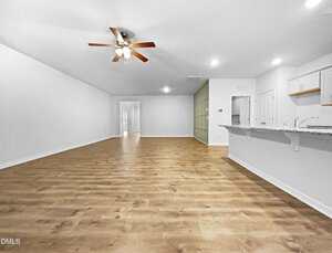 Spacious, empty living area with wood flooring, white walls, ceiling fan with lights, built-in shelves along one wall, and an open kitchen with granite countertops and white cabinets.