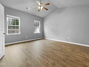An empty room with light gray walls, wood flooring, two windows, a ceiling fan with lights, and a closed white door. The room has a vaulted ceiling and receives natural light from the windows.
