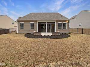 A single-story tan house with a gray roof, fenced backyard, and a screened-in porch. The yard has brown grass, a circular mulched area with stone edging, and is bordered by neighboring houses.