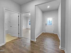 A hallway with wood flooring leads to a white front door with a decorative glass panel. Two open doorways on the left reveal adjacent rooms with light-colored flooring and windows. Walls are painted light gray.