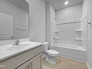 A clean, modern bathroom with a white vanity and sink, a toilet, and a combination bathtub and shower with built-in shelves, all set against light-colored walls and flooring.