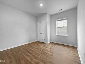 An empty room with light-colored walls, a wood floor, a closed double-door closet, a single window with blinds, and a ceiling light. The room appears clean and well-lit.