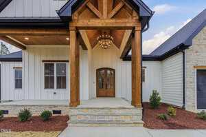 The image shows the front entrance of a house with a wooden double door, covered porch supported by thick wooden beams, stone steps, white siding, and minimal landscaping with small shrubs.