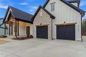 A modern house with light-colored siding and stone accents, featuring two black garage doors, a covered front porch with wooden beams, a concrete driveway, and minimal landscaping.