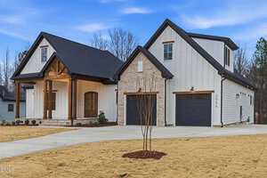 A modern two-story house with light-colored siding, stone accents, dark trim, and a double garage. The front yard has a bare lawn with a small tree, set against a backdrop of trees and a clear sky.