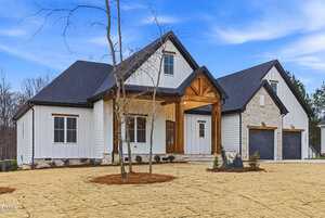 A modern farmhouse with white siding, black roof, wood accents, and stone detailing features a covered front porch, large windows, and a double garage. Young trees and fresh mulch are in the front yard.
