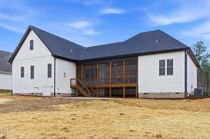 A modern house with white siding and a dark shingle roof. The back features a large screened porch with wooden steps, several windows, and a bare yard covered with straw. The sky is clear with a few clouds.
