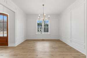 A bright, empty room with white paneled walls, light wood flooring, a large window showing trees outside, and a modern chandelier hanging from the ceiling. An arched doorway is visible on the left side.