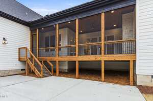 A screened-in wooden porch with black railings and steps leads to a concrete patio. The porch is elevated with open space underneath, attached to a white house with stone accents.
