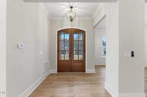 A foyer with light wood flooring, neutral-colored walls, a modern light fixture, and a double wooden front door with arched glass panels, allowing a view of the outdoors.