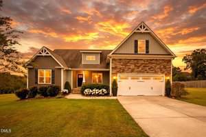 A modern suburban house at 1033 Butterfly Circle in Wake Forest features gray siding, stone accents, and a white double garage on a manicured lawn, set against dramatic orange sunset clouds.