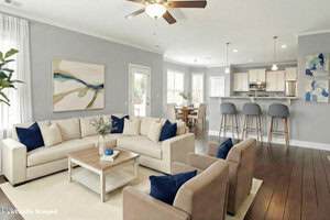 A modern open-plan living room with beige sofas, blue accent pillows, a wooden coffee table, bar stools at a kitchen counter, wall art, and large windows letting in natural light.