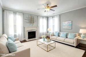 A bright living room with two white sofas, blue accent pillows, a white fireplace, a glass coffee table, two lamps, light curtains on tall windows, and modern wall art above the mantel and sofa.