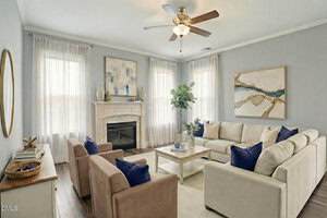 A living room with light gray walls, a ceiling fan, beige sofas and chairs with navy blue pillows, a fireplace, two large abstract paintings, wood flooring, and tall windows with sheer curtains.