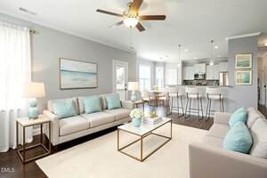 A modern living room with beige sofas, light blue pillows, a glass coffee table, a rug, and wall art. In the background, there is a kitchen with barstools and pendant lights, and a dining area with a table and chairs.