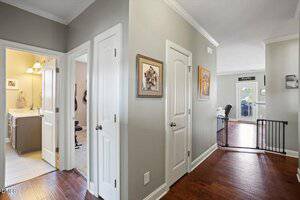 A hallway with wooden floors and light gray walls features white doors on either side. One room with a sink and mirror is visible on the left, and a spacious living area is straight ahead past a black baby gate.