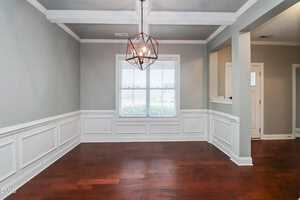A dining room with gray walls, white wainscoting, and a modern geometric light fixture. The room has a large window with white blinds and dark wood flooring. An entryway is visible to the right.