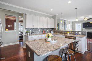 Modern kitchen with white cabinets, stainless steel appliances, granite countertops, a large island with bar stools, and a backsplash of gray tiles. A tray with flowers and decor sits on the island. Dining area visible in the background.