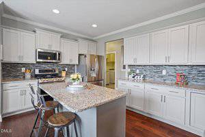 Modern kitchen with white cabinets, stainless steel appliances, a granite island with three wooden stools, and a mosaic tile backsplash. A tray with flowers sits on the island, and "Eat" decor appears on the counter.