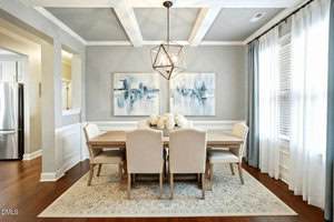 A dining room with a wooden table, six upholstered chairs, a geometric chandelier, two abstract paintings on the wall, a large window with sheer curtains, and a patterned rug on a hardwood floor.