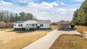 A single-story white house with a front porch sits on a large lawn, with a detached garage and a long driveway leading to both buildings. Trees border the property under a partly cloudy sky.