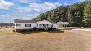 A single-story white house with a covered front porch and attached carport sits on a large, open lawn bordered by tall trees under a partly cloudy sky.