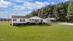 Charming single-story white house at 4577 Hight Road in Oxford, featuring a covered porch, attached carport, and a large front lawn beside tall pine trees under a blue sky with scattered clouds.