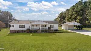 A single-story white house with a front porch and attached brick foundation sits on a large, well-maintained green lawn. A detached carport with a driveway is on the right, and trees border the property in the background.
