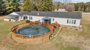 Aerial view of a single-story house with a covered back porch, above-ground pool with a green cover, and a wooden deck. The property is surrounded by grass, trees, and a small outbuilding in the background.