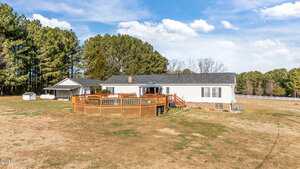 A single-story white house with a dark roof sits on a large grassy lot. There is an above-ground pool with a wooden deck in the backyard, surrounded by trees and open space under a blue sky with scattered clouds.