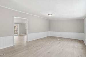An empty room with light wood floors, beige upper walls, white wainscoting, and a ceiling light fixture. An open doorway leads to another room, and a window is visible in the adjacent space.