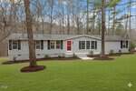Charming single-story gray house at 1613 Hunters Trail in Creedmoor, featuring black shutters and a red front door, set on a well-kept lawn with mulch beds and surrounded by tall trees under a blue sky with scattered clouds.