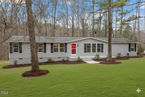 Charming single-story gray house at 1613 Hunters Trail in Creedmoor, featuring black shutters and a red front door, set on a well-kept lawn with mulch beds and surrounded by tall trees under a blue sky with scattered clouds.