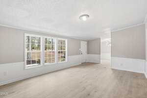A bright, empty living room with light wood flooring, beige walls, white trim, three large windows, and a white front door. Sunlight fills the room, and a dining area is visible in the background.