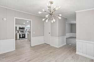 A modern dining area with light wood flooring, beige walls, and white wainscoting, features a sputnik-style chandelier. An open doorway leads to a kitchen with stainless steel appliances.